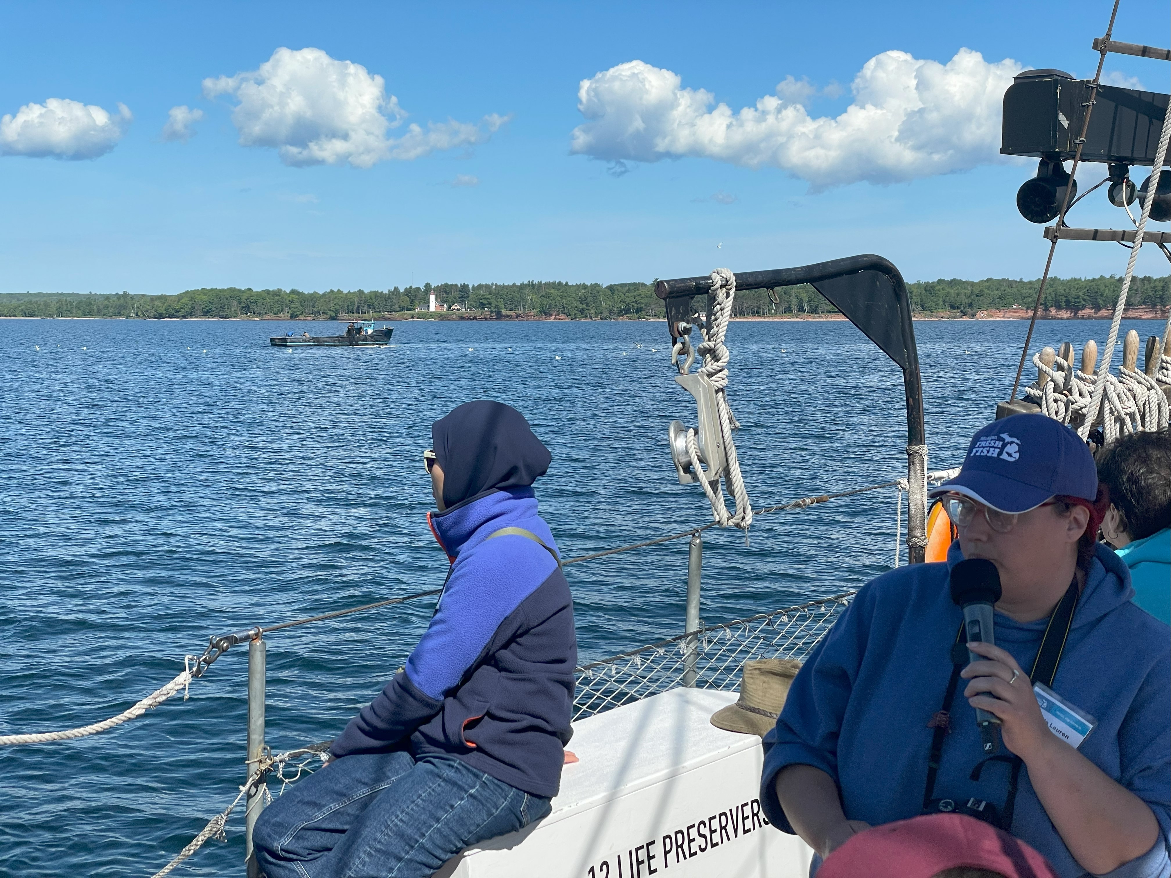 One person sits on the side of a sailboat looking out over the water. Another is talking into a microphone and describing commercial fishing to the listeners.